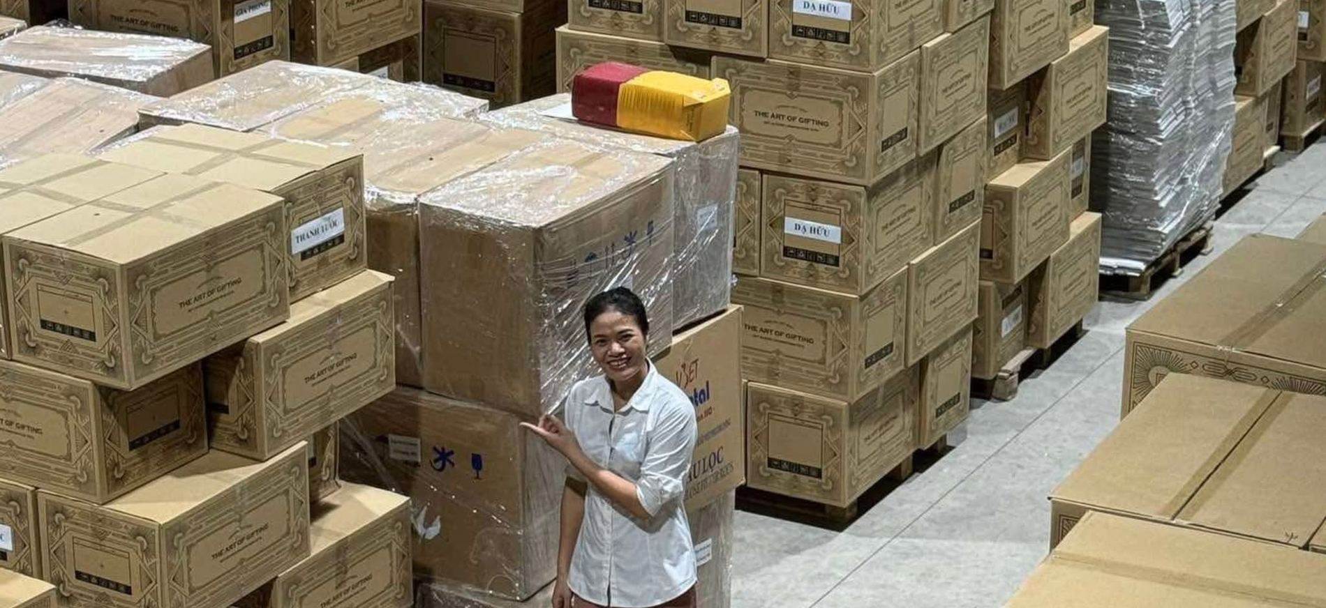 Nhân viên Tetta đứng giữa kho hàng với những pallet thùng hộp quà dán nhãn theo từng mẫu: Gia Phong, Dạ Hữu, Thanh Tước